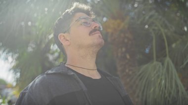 Bıyıklı genç bir İspanyol adam güneşli bir parkta yeşil alanların arasında rahatlıyor. Huzur ve memnuniyet saçıyor..