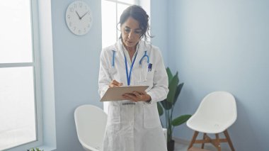 A middle-aged hispanic woman in a white coat writing on a clipboard in a bright hospital room. clipart