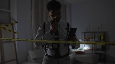 Bir Afro-Amerikan polis dedektifi telsizle konuşur ve kapalı bir suç mahallinde tableti inceler..