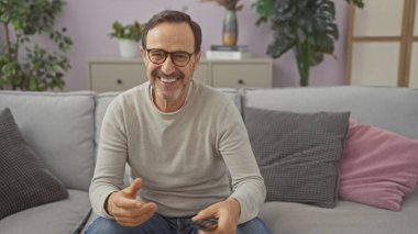 İspanyol olgun bir adam gri bir koltukta oturmuş uzaktan kumandayla gülümsüyor.