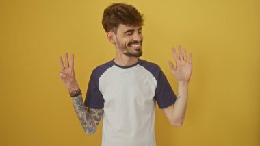 Dövmeli, gülümseyen İspanyol bir adam, sarı arka planda parmaklarıyla sekiz numaralı hareketi yapar..