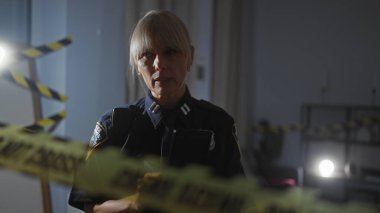 Olgun bir polis memuru dikkatli bir bantla loş ışık alan bir suç mahallini araştırıyor.