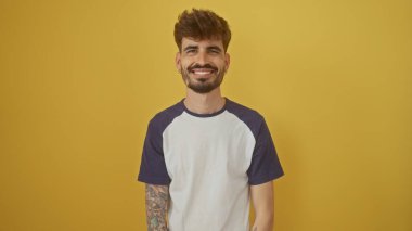 Sakallı ve dövmeli yakışıklı genç adam canlı sarı bir arka plana güvenerek gülümsüyor.