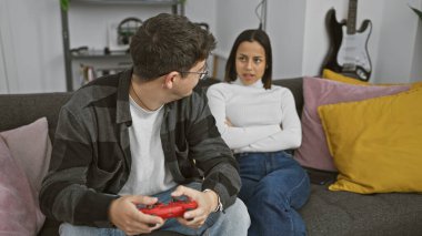 Kollarını kavuşturmuş hoşnutsuz bir kadın, rahat bir oturma odasında video oyunu oynayan bir adamın yanında oturuyor..