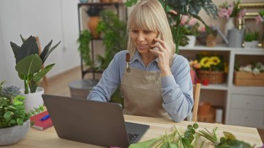 Orta yaşlı, beyaz, önlüklü bir kadın çiçekçi. Dükkandaki bir telefon görüşmesi ve dizüstü bilgisayarda birden fazla iş yapıyor..