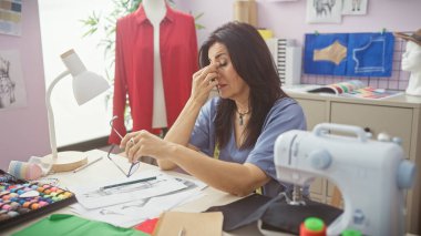 Olgun İspanyol kadın, renkli bir terzi atölyesinde dikiş makinesi, çizimler ve tekstil ile yoruluyor.