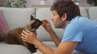 İspanyol bir adam, sıcak bir oturma odasındaki kanepede siyam kedisiyle sevgiyle etkileşim kurar.