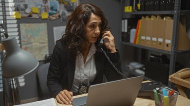 Orta yaşlı bir kadın dedektif ofisinde masasında çalışıyor, telefonla konuşuyor ve dizüstü bilgisayar kullanıyor.