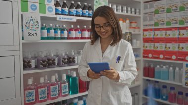 Genç, çekici, esmer bir kadın eczanede beyaz önlük giyiyor ve tablet kullanıyor. Etrafı çeşitli ilaç ürünleriyle çevrili..