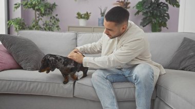 Yakışıklı, genç, İspanyol bir adam rahat bir evin içinde, kanepede dachshund besliyor..