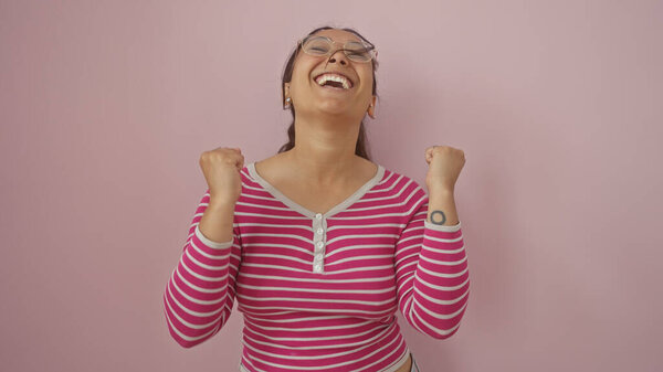 Happy young hispanic woman with glasses celebrating in front of a pink wall