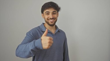 Sakallı yakışıklı genç adam beyaz bir arkaplan karşısında başparmak kaldırıyor.