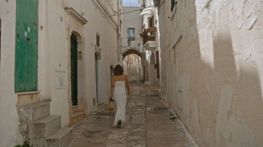Beyaz elbiseli genç bir İspanyol kadın. Ostuni, Puglia, İtalya 'nın eski sokaklarında yürüyor. Arka planda kırsal bir duvar ve yeşil bir kapı var..
