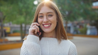 Gülümseyen beyaz saçlı kadın güneşli bir açık hava parkında neşeli bir telefon görüşmesi yapıyor.