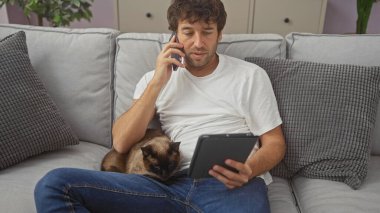 İspanyol adam rahat bir dairede siyam kedisiyle kanepede dinlenirken telefon ve tabletle birden fazla iş yapıyor..