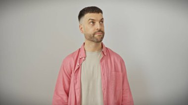 Beyaz bir arkaplanı olan, günlük pembe gömlekli, sakallı yakışıklı, genç, İspanyol bir adam dalgın görünüyor.