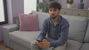 Modern bir apartman dairesinde oturan sakallı yakışıklı Arap adam akıllı telefon kullanıyor.