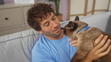 İspanyol bir adam sıcak bir ev ortamında bir siyam kedisini şefkatle tutuyor, özen ve arkadaşlık gösteriyor..