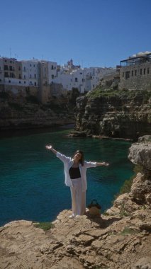 Genç bir İspanyol kadın, polignano 'da denize bakan kayalık bir uçurumda neşeyle duruyor. Bir kısrak, puglia, İtalya, arka planda beyaz binalar..