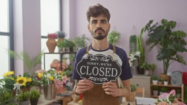 A young bearded man holds a 'closed' sign in a floral shop filled with plants and flowers. clipart