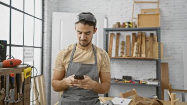 Sakallı İspanyol adam marangozluk atölyesinde ahşap ve aletlerle dolu akıllı telefon kullanıyor..