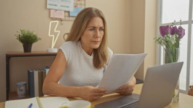 Masasında dizüstü bilgisayarı, çiçekleri ve kahvesiyle evrak okuyan olgun bir kadın.