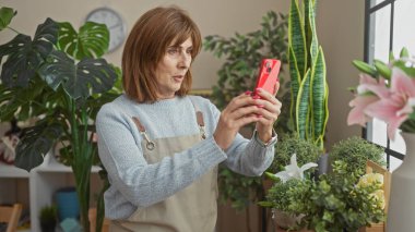 Bir kadın akıllı telefonunu kullanarak parlak bir çiçekçide çiçeklerin fotoğraflarını çekmekten hoşlanır..