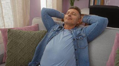 Orta yaşlı bir adam yastıklı ve sakin bir ortamda elleri başının arkasında kanepede dinleniyor.