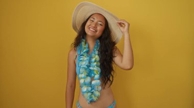 Yaz şapkası takan genç bir kadın ve çiçek kolyesi izole edilmiş sarı bir arka plana gülümsüyor..