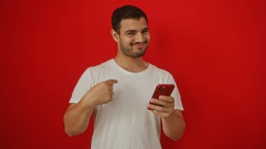 Beyaz tişörtlü, akıllı telefon kullanan ve kırmızı arka planda kendini gösteren İspanyol bir genç.