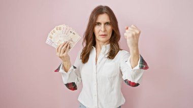 Kısa saçlı, kuşkucu orta yaşlı bir kadın pembe arka planda Türk lirası tutuyor.
