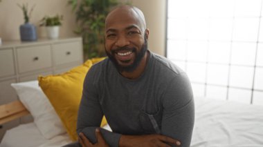 Sakallı genç adam rahat yatak odasında, çiçekli ve yastıklı bir şekilde oturuyor, evde güvenle gülümsüyor.