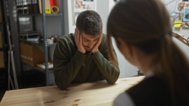 Distressed man interrogated by woman in a cluttered investigation room, suggesting a suspenseful encounter. clipart