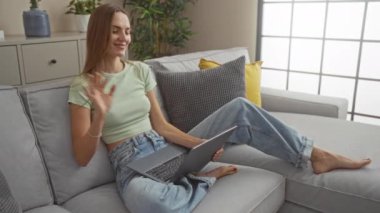 Dizüstü bilgisayarda konuşan bir kadın modern bir oturma odasında arka planda yastıklar ve bitkiler olan bir koltukta oturuyor..
