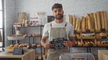 Fırındaki genç adam elinde açık bir tabela ile arka plandaki raflarda çeşitli ekmek ve hamur işleri sergiliyor.