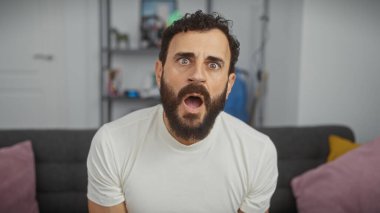 Oturma odasında beyaz giysili, ağzı açık, gözleri açık, ağzı açık, şaşkın sakallı bir adam..