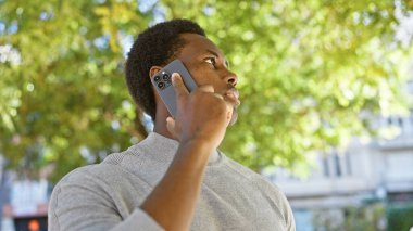 Açık havada, yeşil yapraklarla çevrili akıllı telefondan konuşan Afrikalı genç bir Amerikalı..