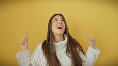 Neşeli gülümseyen İspanyol kadın, beyaz bir süveter giyen, hayat dolu sarı bir arka planda izole edilmiş, neşeyle yukarıya bakıyor..