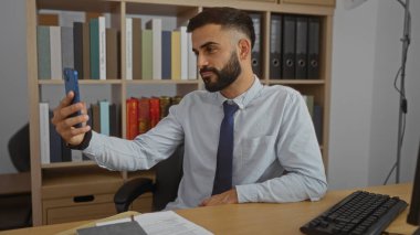 Ofis odasında selfie çeken sakallı genç İspanyol bir adam. Arka planda bilgisayar ve kitaplıkla masada oturuyor.