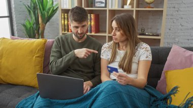 Endişeli bir kadın rahat bir oturma odasında kredi kartı tutarken bir erkek dizüstü bilgisayarı işaret eder.