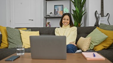 Gülümseyen genç İspanyol kadın evinde kanepede dizüstü bilgisayarı, arka planda gitarı, kaktüs neon ışığı ile uzanıyor.