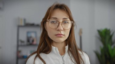 Gözlüklü beyaz bir kadın, iyi aydınlatılmış bir oturma odasında tarafsız bir ifadeyle, sakin ve sade bir şekilde poz veriyor..