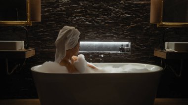 Genç bir kadın, modern, şık bir banyoda sükunetle sarılı köpükle dolu lüks bir küvette dinleniyor..
