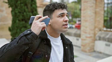 Kıvırcık saçlı yakışıklı adam kalabalık bir caddede telefonundaki sesli mesajı dinliyor..