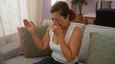 Orta yaşlı bir kadın evde yeşil yastıklarla oturma odasında otururken burnunu kötü kokudan koruyor..