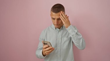 Kısa saçlı ve açık mavi gömlekli genç adam telefonunun pembe arka planıyla karşılaştırılırken stresli görünüyor..