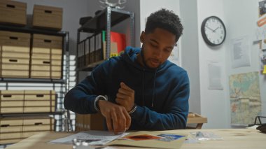 Bir dedektifin ofisindeki ipuçlarını inceleyen Afro-Amerikalı bir adam. Etrafı delil ve dava dosyalarıyla çevrili..