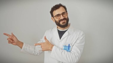 Laboratuvar önlüklü gülümseyen sakallı adam beyaz arka planı işaret ediyor.