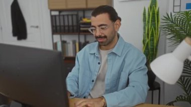 Modern ofiste çalışan bıyıklı genç bir adam gözlük takıyor ve farklı heyecan ifadeleri sergiliyor ve bilgisayardaki görevlerine odaklanıyor.