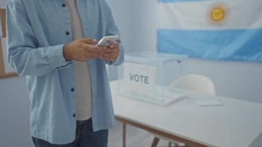 Seçmen kolej odasında akıllı telefon kullanan İspanyol bir adam. Masasında Arjantin bayrağı, afişi ve saydam bir oy sandığı var..